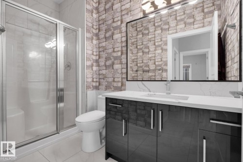 Glass-enclosed shower with chrome fixtures, white tile flooring, white brick-patterned accent wall, vanity with dark high-gloss cabinetry, and a large mirror with a black frame - #105 160 Magrath Rd Nw, Edmonton, AB 