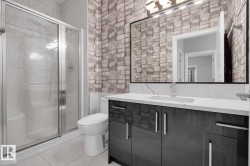 Glass-enclosed shower with chrome fixtures, white tile flooring, white brick-patterned accent wall, vanity with dark high-gloss cabinetry, and a large mirror with a black frame - 