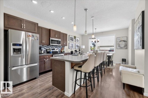 Kitchen featuring wood-finish flooring, stainless steel appliances, dark wood cabinetry, a tiled backsplash, and a breakfast bar with pendant lighting - 2046 Chalmers Way Sw, Edmonton, AB 