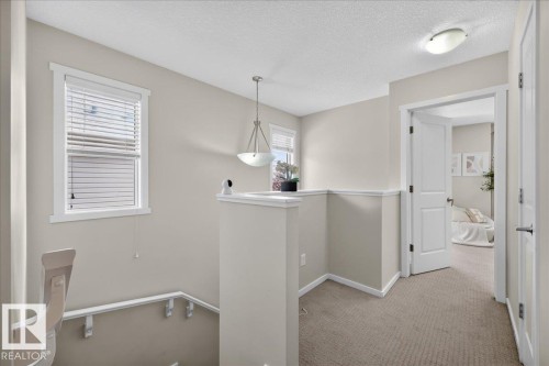 Carpeted hallway with a window featuring blinds, a hanging pendant light, an open doorway, and a flush mount ceiling light - 2046 Chalmers Way Sw, Edmonton, AB 