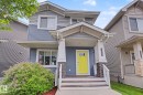 Front elevation featuring blue horizontal siding, decorative gray shake siding, a covered porch with stone-clad pillars, a bright yellow entry door, and a mature flowering shrub - 3343 Weidle Way Sw, Edmonton, AB 
