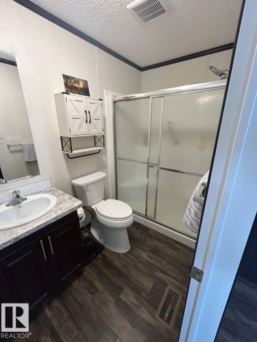 Dark wood-finish flooring, white vanity with granite-patterned countertop, sliding glass shower enclosure, white wall cabinetry with dark hardware, and dark crown molding - 63 Willow Park Estates, Leduc, AB 