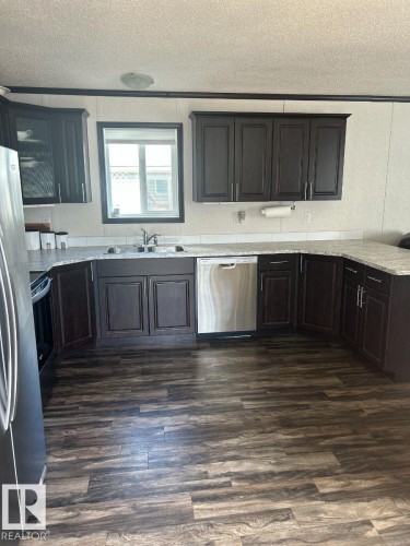 U-shaped kitchen featuring dark wood-finish cabinetry, light-toned countertops, wood-finish flooring, a stainless steel dishwasher, and a window above the sink - 63 Willow Park Estates, Leduc, AB 