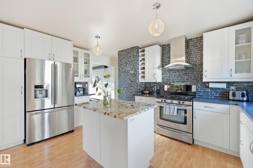 Kitchen featuring white cabinetry, stainless steel appliances, a central island with a stone countertop, a stone accent wall, and wood-finish flooring - 3523 106 Street Nw, Edmonton, AB 