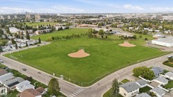 Expansive green space featuring multiple baseball diamonds and soccer fields, surrounded by residential properties and a distant city skyline - 