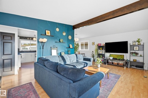 Open-concept living area featuring a prominent exposed wood beam, wood-finish flooring, a vibrant accent wall, and a large window providing natural light - 3523 106 Street Nw, Edmonton, AB 