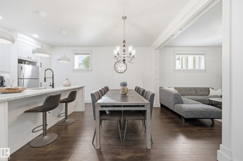 Open-concept layout featuring wood-finish flooring, white cabinetry, a kitchen island with seating, stainless steel appliances, and an elegant chandelier - 1961 Adamson Terrace Sw, Edmonton, AB 