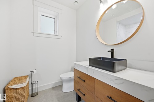 Modern vanity featuring wood-finish cabinetry, a rectangular vessel sink, a stone-finish countertop, and a round wall-mounted mirror - 1961 Adamson Terrace Sw, Edmonton, AB 