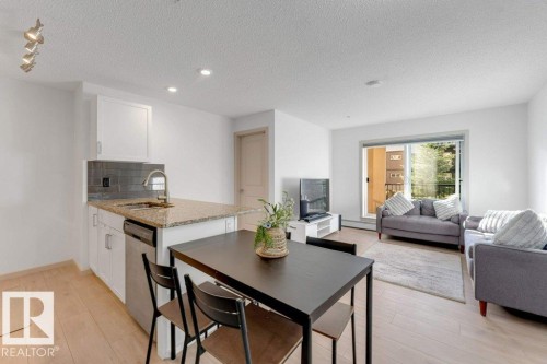Open-concept living area with wood-finish flooring, white cabinetry, a kitchen island with stone countertop, and a sliding glass door providing access to an exterior balcony - #224 390 Windermere Rd Nw, Edmonton, AB 