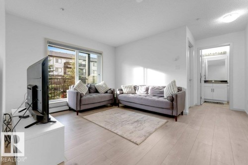 Light wood-finish flooring, sliding glass door leading to a balcony, and a white vanity with a dark countertop and mirror - #224 390 Windermere Rd Nw, Edmonton, AB 