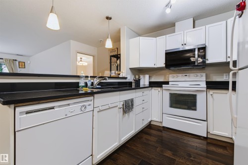Kitchen featuring white cabinetry, dark wood-finish flooring, dark countertops, and stainless steel sink with a goose-neck faucet - #155 2096 Blackmud Creek Dr Sw, Edmonton, AB 