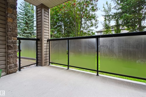 Concrete patio featuring a glass-panel railing with black metal posts, stone veneer accent column, and horizontal siding - #155 2096 Blackmud Creek Dr Sw, Edmonton, AB 
