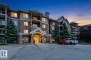 Multi-story building with stone and siding exterior, prominent entrance with overhead lighting, multiple balconies, and mature evergreen landscaping - #155 2096 Blackmud Creek Dr Sw, Edmonton, AB 