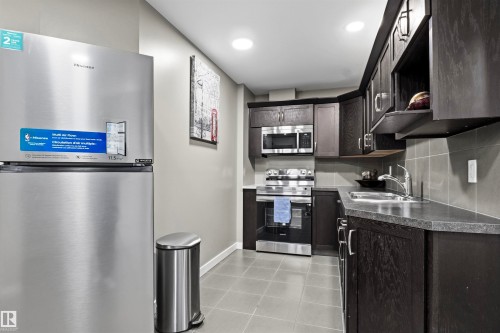 Galley kitchen featuring dark wood-finish cabinetry, stainless steel appliances, and light gray tile flooring - 8515 19 Avenue Sw, Edmonton, AB 