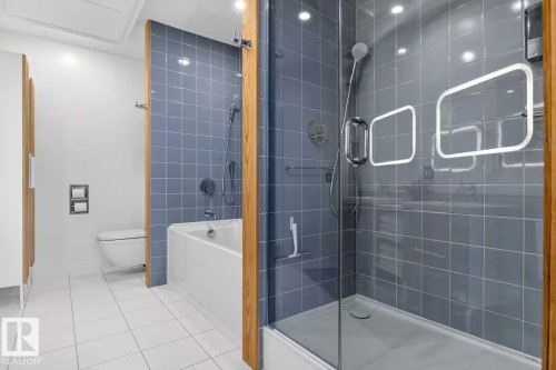Bathroom featuring a glass-enclosed shower, a built-in tub, blue wall tile, white floor tile, and a wall-mounted toilet - #501 10105 109 St Nw, Edmonton, AB 