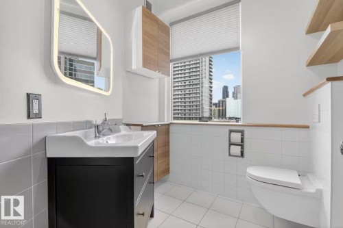 White tile flooring, wall-mounted toilet, floating wood-finish shelving, vanity with integrated sink, and an illuminated mirror - #501 10105 109 St Nw, Edmonton, AB 