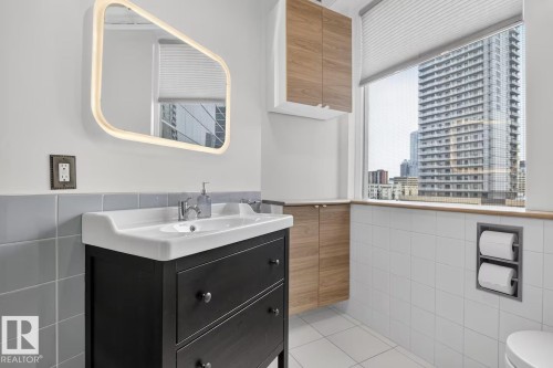 Integrated lighting mirror, dark wood-finish vanity with white basin, wood-finish wall-mounted cabinetry, white tiled flooring, and a large window - #501 10105 109 St Nw, Edmonton, AB 