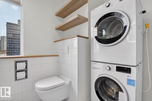 Compact laundry area featuring a stacked washer and dryer, white tiled wall, and wood-finish shelving - #501 10105 109 St Nw, Edmonton, AB 