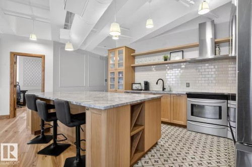 Expansive kitchen island with a light-toned countertop, natural wood-finish cabinetry, stainless steel oven, white subway tile backsplash, and decorative floor tiling - #501 10105 109 St Nw, Edmonton, AB 