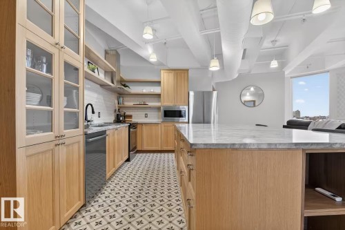 Expansive kitchen island with a stone countertop, light wood cabinetry, patterned floor tiling, exposed ceiling ductwork, and pendant lighting fixtures - #501 10105 109 St Nw, Edmonton, AB 