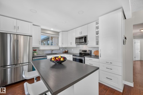 Kitchen featuring white cabinetry, stainless steel appliances, dark countertops, a mosaic tile backsplash, and wood-finish flooring - 7704 13 Avenue Nw, Edmonton, AB 