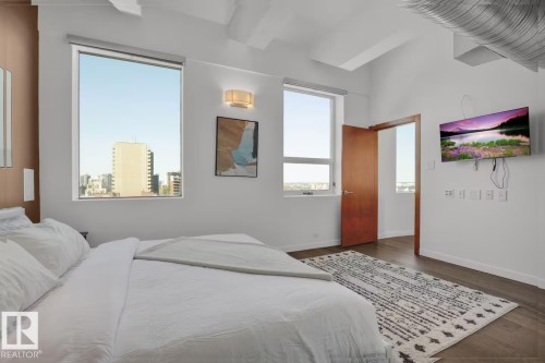 High ceilings with exposed ductwork, wood-finish flooring, two large windows, a solid core wood door, and a wall-mounted flat-screen television - #701 10105 109 St Nw, Edmonton, AB 