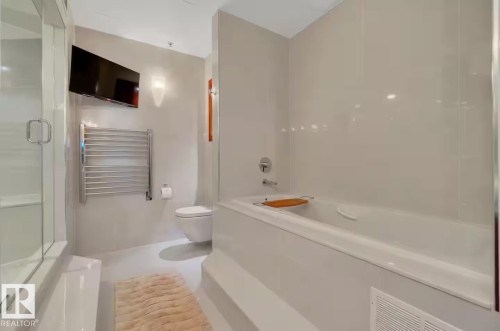 Built-in tub with chrome fixtures, glass-enclosed shower, wall-mounted television, chrome towel warmer, and wall-mounted toilet - #701 10105 109 St Nw, Edmonton, AB 