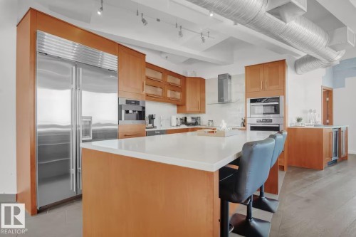 Kitchen featuring wood-finish cabinetry, stainless steel appliances, a center island with a light-toned countertop, and exposed ductwork - #701 10105 109 St Nw, Edmonton, AB 