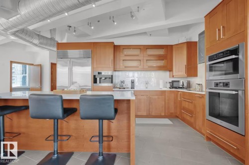 Kitchen featuring light wood cabinetry, stainless steel appliances, a white tile backsplash, a large island with a white countertop, and exposed ductwork - #701 10105 109 St Nw, Edmonton, AB 