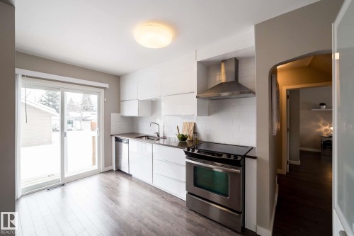 Modern kitchen with wood-finish flooring, white cabinetry, stainless steel appliances, and a sliding glass door - 12228 134 Street Nw, Edmonton, AB 