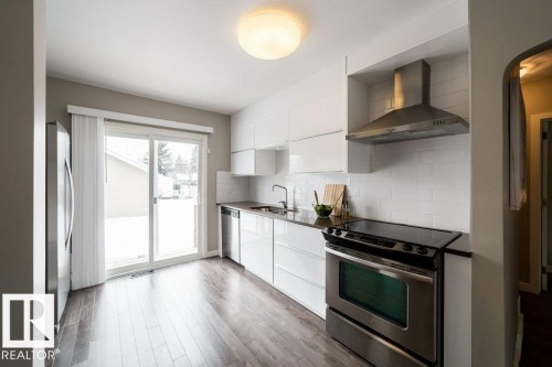 Kitchen featuring stainless steel appliances, white flat-panel cabinetry, dark countertops, white subway tile backsplash, and wood-finish flooring - 12228 134 Street Nw, Edmonton, AB 