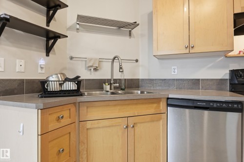 Stainless steel dishwasher, double basin sink with a high-arc faucet, light wood-finish cabinetry, and dark gray countertop surfaces - #85 150 Edwards Dr Sw, Edmonton, AB 