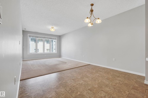 Expansive room featuring a large window, mixed flooring with carpet and tile, a brass-tone chandelier, and a flush-mount ceiling light - #215 9820 165 St Nw, Edmonton, AB 