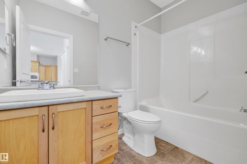 Bathroom featuring a single vanity with wood cabinetry, a rectangular sink, a large mirror, a toilet, and a white bathtub with a shower surround - #215 9820 165 St Nw, Edmonton, AB 