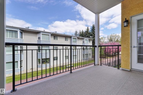 Covered balcony with a textured concrete surface, black metal railings, and an exterior wall featuring a stucco finish - #215 9820 165 St Nw, Edmonton, AB 