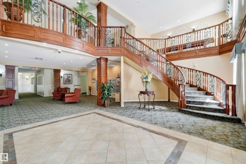 Expansive foyer featuring a grand curved staircase with wrought iron and wood railings, tile flooring with decorative inlay, patterned carpet, and built-in lighting - #215 9820 165 St Nw, Edmonton, AB 