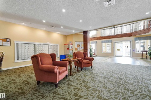 Spacious common area featuring patterned carpeting, recessed lighting, a built-in mail center, and an upper-level balcony with decorative railing - #215 9820 165 St Nw, Edmonton, AB 
