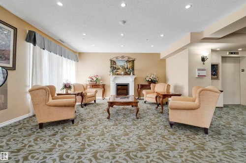 Spacious common area featuring patterned carpeting, recessed ceiling lighting, a decorative fireplace, and an elevator - #215 9820 165 St Nw, Edmonton, AB 
