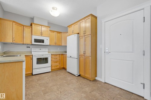 Kitchen featuring light wood-finish cabinetry, white appliances, neutral countertops, and patterned tile flooring - #215 9820 165 St Nw, Edmonton, AB 