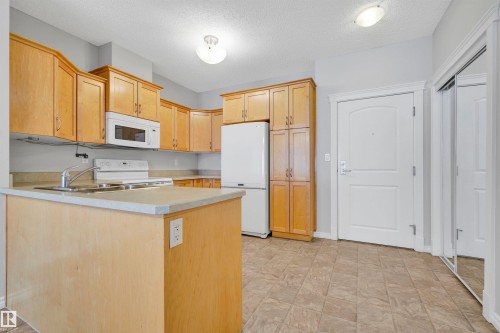 Kitchen featuring wood-finish cabinetry, white appliances, and a peninsula with integrated sink - #215 9820 165 St Nw, Edmonton, AB 