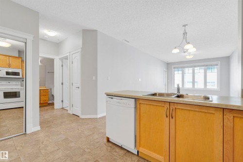Kitchen featuring wood-finish cabinetry, a stainless steel double basin sink, and a white dishwasher - #215 9820 165 St Nw, Edmonton, AB 