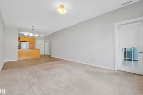 Carpeted living area with light gray wall paint, kitchen with wood-finish cabinetry, stainless steel refrigerator, and pendant lighting - #215 9820 165 St Nw, Edmonton, AB 