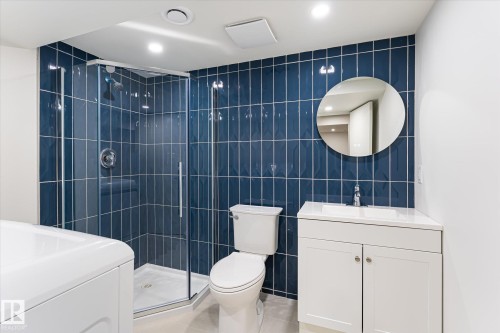 Glass-enclosed shower with blue tile surround, white vanity with integrated sink, modern circular mirror, and recessed lighting - #3 18010 98 Av Nw, Edmonton, AB 