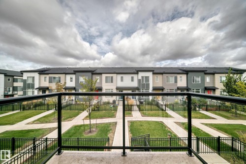 Black metal and glass panel railing with a concrete deck, overlooking a structured green space with walkways and multi-story residential buildings featuring various exterior finishes - #56 804 Welsh Dr Sw, Edmonton, AB 
