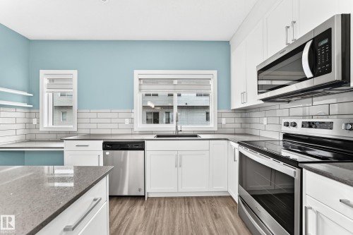 Kitchen featuring white cabinetry, stainless steel appliances, dark countertops, white subway tile backsplash, and wood-finish flooring - #56 804 Welsh Dr Sw, Edmonton, AB 