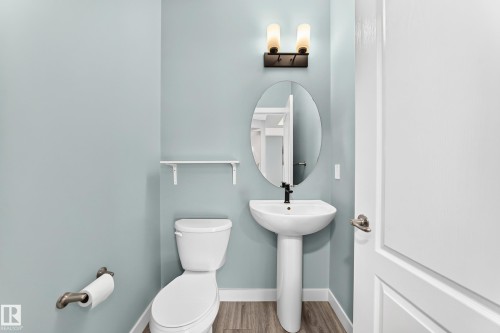 Pedestal sink with a matte black faucet, oval mirror, wall-mounted sconce lighting, and wood-finish flooring - #56 804 Welsh Dr Sw, Edmonton, AB 