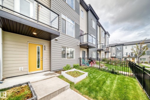 Modern multi-level exterior with horizontal siding, a vibrant entry door, glass panel balcony railings, a manicured lawn, and black metal fencing - #56 804 Welsh Dr Sw, Edmonton, AB 