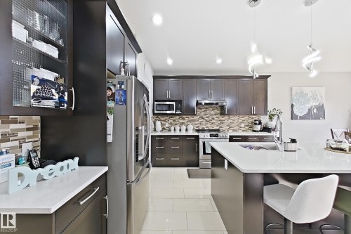 Well-appointed kitchen featuring dark wood-finish cabinetry, stainless steel appliances, a mosaic tile backsplash, and an island with a white countertop - 144 52 Street Sw, Edmonton, AB 