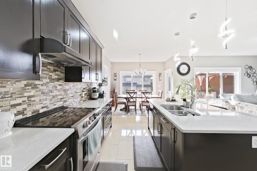 Kitchen featuring dark wood cabinetry, a mosaic tile backsplash, stainless steel appliances, a center island with a double sink, and pendant lighting - 144 52 Street Sw, Edmonton, AB 
