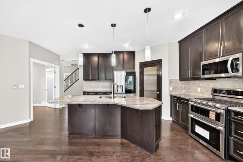 Kitchen featuring a central island with an undermount sink, pendant lighting, stainless steel appliances, dark wood cabinetry, and wood-finish flooring - 3329 West Court Nw, Edmonton, AB 
